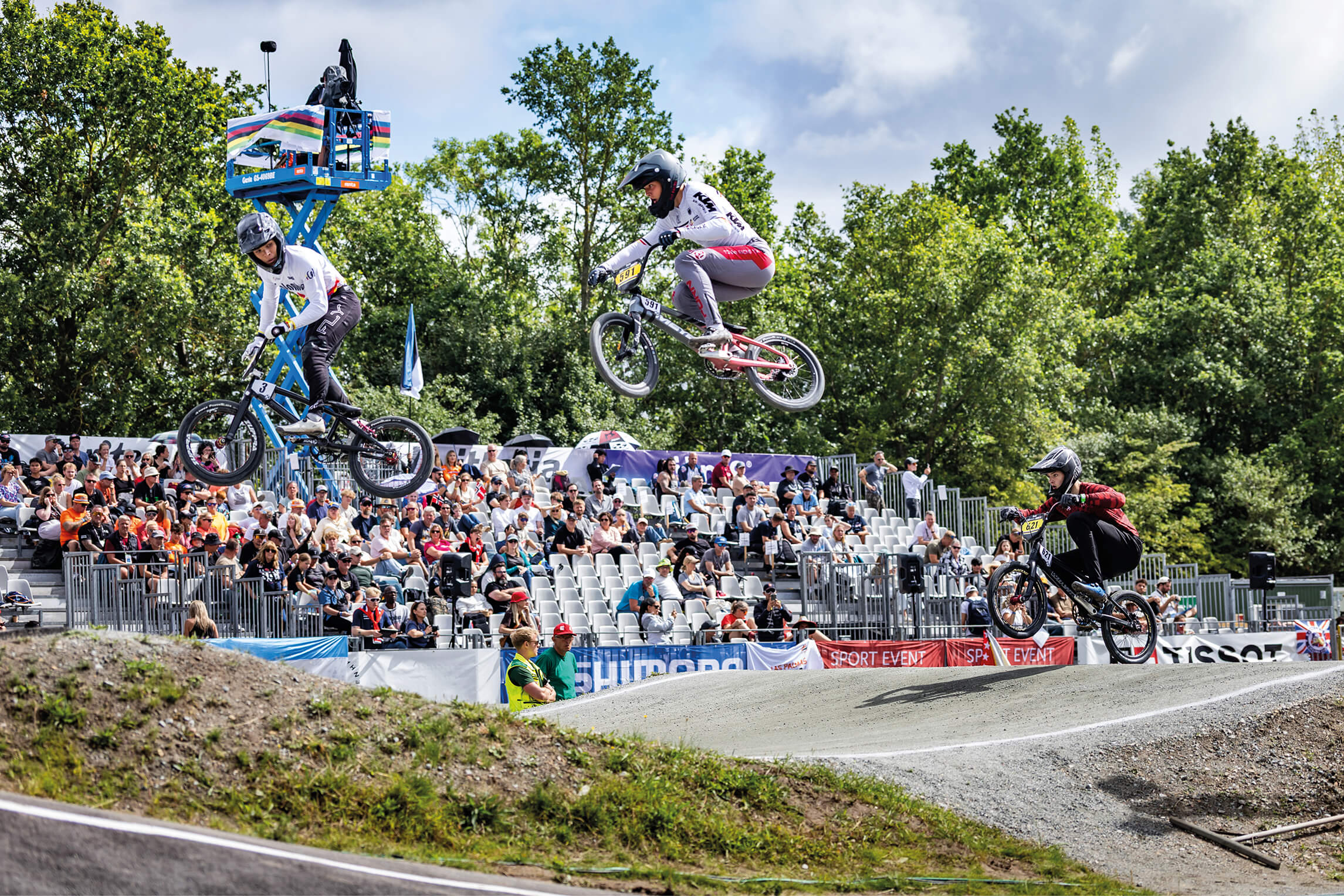 Drei BMX-Racer springen über ein Hindernis, mittig Louis Romeyke im weißen Deutschlandtrikot vor Zuschauertribüne