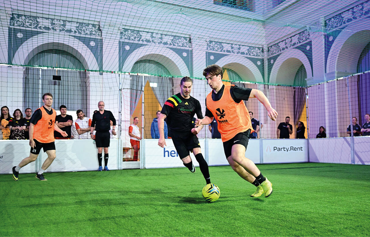 Drei Fußballspieler im Indoor-Spiel mit Schiedsrichter und Zuschauern im Börsensaal der Handelskammer Hamburg